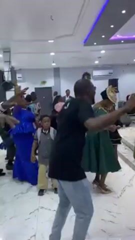 The video captures a group of people dancing and celebrating in a brightly lit, modern indoor space with patterned tile flooring. Most of the individuals are African, dressed in a variety of clothing, including traditional African prints, dresses, shirts, and jeans. Some are holding their hands in the air, swaying, and generally seem joyful. The room appears to be a reception hall or large community room, with white walls, recessed lighting, and decorative ceiling panels. There is also a sound system visible in the background.