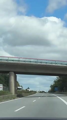 A vehicle is driving on a highway. The sky is cloudy. There is a concrete overpass above the highway with a red and green railing. On the side of the highway, there are trees, some signage, and a power plant in the distance. Another vehicle is visible further ahead on the highway.