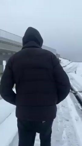A person in a black jacket walks along a snowy embankment towards a partially constructed bridge in a snowy, overcast environment. The bridge spans a body of water, partly covered with ice and snow. The view then pans to reveal more of the unfinished bridge and the winter landscape. The water reflects the grey sky, adding to the bleak atmosphere.