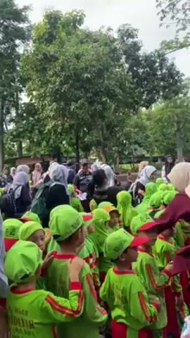 A group of children wearing matching green and red outfits are gathered outdoors, apparently with their teachers and other adults. The children are wearing green caps and long-sleeved shirts with red accents and an embroidered logo, and some are hugging each other. The adults accompanying them appear to be primarily female, with several wearing hijabs. They are standing among trees in an outdoor area.
