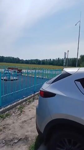 A silver SUV is parked in front of a fenced-in playground. The playground has blue rubber surfacing, a blue and turquoise fence, and several pieces of equipment, including a slide and a merry-go-round. Two adults and a child are visible in the playground. In the background, there is a field of trees, streetlights, and a clear sky. The weather appears to be sunny.