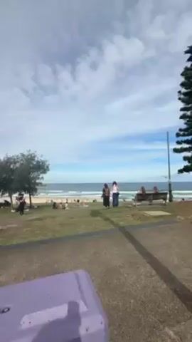 The video begins with a shot of a beach scene with a grassy area leading to the sand and ocean. There are people sunbathing and walking around. The camera then quickly shifts to a view of buildings and a person wearing a helmet riding a scooter. The view becomes blurry and angled upward toward the sky. The camera angle moves down, focusing on the rider's bare feet on the scooter's footrest. The scooter's keys are visible in the ignition.
