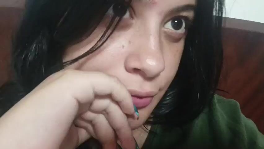 A young woman with dark hair, dark eyes, and a mole on her forehead is filmed in a close-up shot, wearing a dark green top. Her left thumb with a green-painted nail is initially near her chin. She looks around, shifting her gaze between the camera and points slightly above and to the side. At one point, she uses her right hand to adjust her hair. Her lips are slightly parted, and she makes subtle mouth movements. The background is blurred and appears to be indoors.