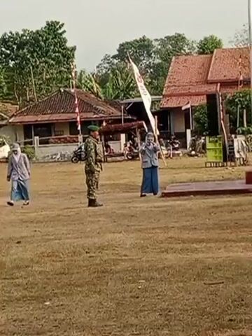 The video shows an outdoor scene with a dirt field in the foreground. Two people are visible. On the left, a person in camouflage military uniform stands at attention facing to the right. On the right, a person wearing a hijab and a light-colored jacket over a blue outfit stands facing the military person. This person holds a banner with some writing and a red symbol on it, and appears to be speaking or leading an event. Behind them, a flagpole stands tall, and a building with a red tiled roof is visible. There are also trees and other structures in the background. The lighting suggests it is daytime.