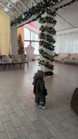 A short, eye-level shot opens on an empty, decorated room. The camera follows a small child in a green jumpsuit and hat, as they walk across the tile floor. The room is brightly lit and sparsely furnished. A Christmas tree, and garland wrapping around a pole are the most prominent features. Large windows let in natural light. The child stops and looks toward the camera briefly before the video ends.