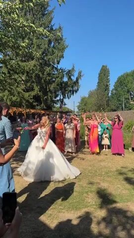 A bride in a white wedding dress with a voluminous skirt throws a bouquet in a grassy outdoor area. She is facing away from the camera, towards a group of women dressed in various colorful dresses, some of whom are reaching out to catch the bouquet. A man in a blue patterned shirt and jeans is standing to the left of the frame, facing the bride, and appears to be filming her with a smartphone. Another person is partially visible in the foreground to the left, also holding a smartphone. The scene is lit by bright sunlight, with trees and a wooden structure in the background, suggesting a wedding reception or ceremony in a garden setting. The sky is clear and blue.