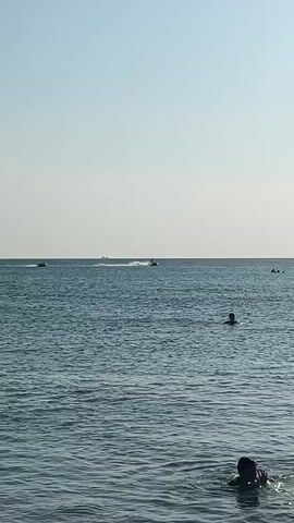 The video shows a view of the sea on a sunny day. The sky is clear and blue, and the water is calm with gentle waves. Several people are visible in the water, some swimming and others enjoying water sports. In the distance, a jet ski is seen speeding across the water, creating a white wake. There are also a couple of distant boats on the horizon. The overall atmosphere is one of relaxation and recreation.