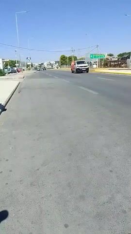 A video taken from a low angle shows a street scene. A white van with orange accents is driving down the road from right to left. Several other cars are also visible on the road. A traffic light is visible in the distance. There are trees and buildings along the side of the road. The sky is clear and blue.