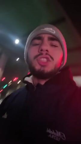 Two young men stand side-by-side outdoors at night. The man on the right, who appears to be filming, wears a gray Nike beanie and a dark jacket with white lettering. He has a beard and mustache, and his mouth is open as if he is talking or singing. The other man wears a tan beanie and a shiny black puffer jacket. He stands slightly behind and to the left of the first man, holding what appears to be a small, lit object in his hand. Behind them is a bus schedule kiosk with digital display screens. Streetlights and the glow from inside a nearby building illuminate the scene. The camera is handheld and moves slightly.