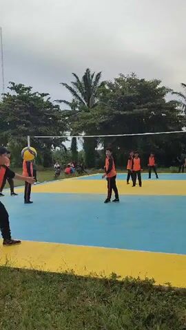 A group of young people wearing orange vests over dark shirts and pants are playing volleyball on an outdoor court. The court is painted blue with yellow borders. Palm trees and other foliage are visible in the background under a cloudy sky. The ball is in the air, and several players are positioned to hit it. One player in the foreground is facing away from the camera. Another player is in the middle of the court, preparing to hit the ball. Several other players are on both sides of the net.