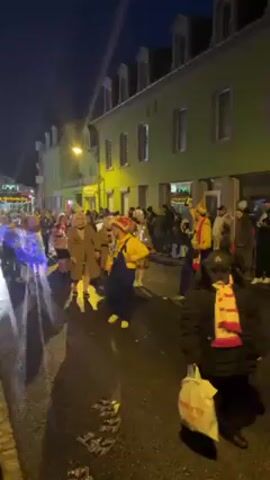The video shows a nighttime parade on a street in a town. The buildings lining the street are two-story and painted green. Streetlights illuminate the scene, and there is a light-up sign in the distance. People are walking in the parade, wearing costumes and holding instruments. Some are wearing light-up clothing. There are spectators on the sidewalks. The street surface is wet, and there are objects scattered on the ground.
