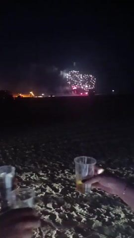 The video begins with a dark scene. In the foreground, several hands are holding plastic cups filled with a light-colored liquid, presumably toasting. The cups are on a sandy surface. In the background, there are distant lights and fireworks exploding in the night sky. The fireworks are the primary light source in the background, illuminating the scene with bursts of red and white light. The video appears to be taken from a low angle, looking up towards the fireworks. The overall atmosphere suggests a celebration, possibly on a beach or an outdoor venue at night. The focus is on the hands and cups in the foreground, with the fireworks serving as a backdrop.