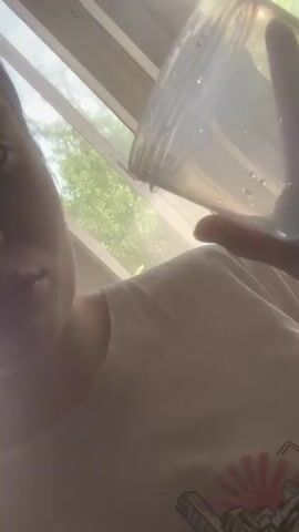 A person holds a clear plastic container in front of the camera. They are wearing a white t-shirt with a graphic design. Through the window behind them, a tall industrial chimney is visible. There are also trees and a building with text on it.