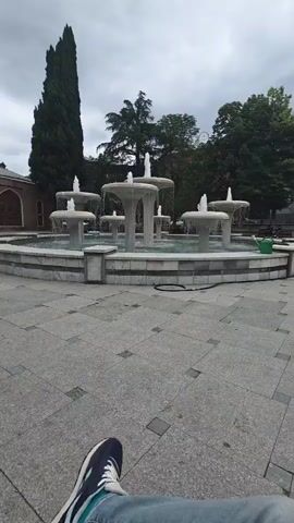The media file shows a fountain in Ganja, Azerbaijan. The fountain has multiple tiers and is surrounded by a tiled plaza. In the foreground, a person's legs and feet are visible, suggesting they are sitting and observing the fountain. The weather appears to be overcast, with a cloudy sky. Lush green trees are visible in the background, adding to the park-like setting. A tall, slender tree stands to the left of the fountain, and a green watering can is placed on the edge of the fountain's basin. The scene evokes a sense of calm and tranquility, possibly a moment of leisure. There is no visible text in the media file.