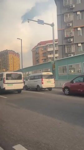 The video shows a view from a car driving on a road. Multiple vehicles are visible, including white vans, a green SUV, and other cars. Buildings and a tall communication tower are present in the background. The sky is cloudy. The vehicles appear to be in traffic or moving slowly on a multi-lane road.