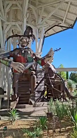 The video shows a static, full shot of a large, colorful metal sculpture of a jester playing a stringed instrument. The jester is seated and appears to be outdoors under a white, wooden canopy structure. To the right of the jester is another, smaller metal sculpture of a person seated with a large wooden wheel. In the background, there is foliage and a clear blue sky. The ground in front of the sculptures is covered with dirt and some plants.