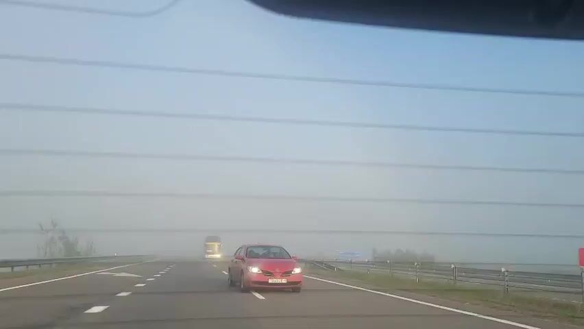 The video shows a view from inside a vehicle traveling on a highway with low visibility due to fog. A red car is visible in the foreground, driving in the same direction. Further ahead, a truck and a white van are seen in the distance, partially obscured by the fog. The road is paved and has white lane markings. The sky is clear and blue above the fog bank.