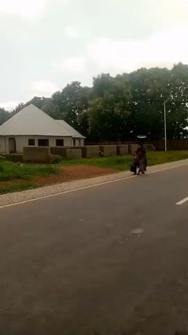 A video clip shows a car driving down a road. The road is paved and has a white line down the middle. On the left side of the road, there is a building and trees. On the right side of the road, there are more trees. The sky is overcast. The video is shaky, indicating it was likely filmed from a moving vehicle.