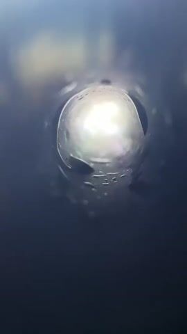 The video begins with a close-up shot of the inside of a dark tube, illuminated by a bright light at the end. A person's hand is then shown holding a clear plastic cup with a black lid and a black straw. The cup contains a reddish-orange liquid and what appear to be fruit pieces. The person is holding the cup and shaking it, causing the contents to swirl. The background is blurred, but appears to be an indoor setting with a table and some items on it. The person's leg, clad in dark pants, is visible in the lower part of the frame. The video cuts between the inside of the tube and the person holding and shaking the cup.