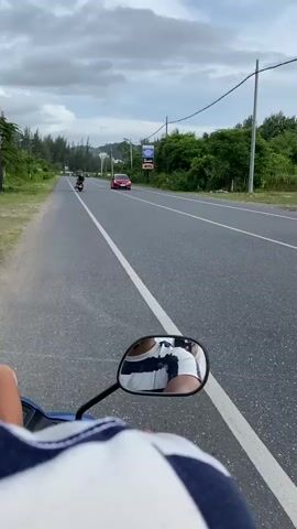 The media file shows a first-person perspective of someone riding a motorcycle on a paved road. In the foreground, the rider's shoulder and arm, clad in a blue and white striped shirt, are visible. A motorcycle's rearview mirror is also prominent, reflecting the rider's torso and the back of their head, which is wearing a striped shirt.

The road stretches ahead, lined with a grassy shoulder on the left and trees on the right. A white line divides the lanes of the asphalt road. Several vehicles are on the road: a red car in the distance, a black SUV approaching, and a few motorcycles further ahead. There are also utility poles and wires visible along the roadside. In the background, lush green hills rise under a cloudy sky, suggesting a somewhat overcast day. There's a billboard on the right side of the road with text that is mostly illegible from this distance.

The scene appears to be set in a rural or semi-rural area, likely in Lamkruet, Indonesia, given the location context. The activity is simply traveling on a motorcycle, likely for transportation or leisure. The overall mood is neutral, focusing on the journey itself. There are no overt expressions of emotion.