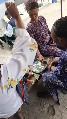Here's a detailed description of the video:

Three women are seated outdoors, engrossed in a game played on a small tray. The woman in the center wears a floral print dress and a headscarf. To her left, another woman is dressed in a white top with yellow flowers and a patterned dark-colored skirt. On the right, the third woman is clad in a blue patterned outfit. They are gathered around what appears to be a makeshift table or low structure. 

In the background, two men are seated at a table playing a different game. One man wears a white shirt and dark baseball cap, while the other is in a maroon shirt. The setting looks like an outdoor public space, possibly a courtyard or a shaded area. The ground is concrete and the area is surrounded by partial structures or tarps. The lighting indicates it is daytime.