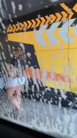 This media file depicts a woman in a car wash. The perspective is from inside the car, looking out through the windshield which is covered in soap suds and water. The woman is visible in the reflection on the windshield, wearing a black top and white shorts. She appears to be looking towards the car wash equipment. The background shows the colorful interior of the car wash, with black, yellow, and orange stripes and chevron patterns. The lighting suggests it is daytime. There is no visible text. The scene captures the sensory experience of being in a car wash, with the suds obscuring the view and creating a dynamic visual effect.