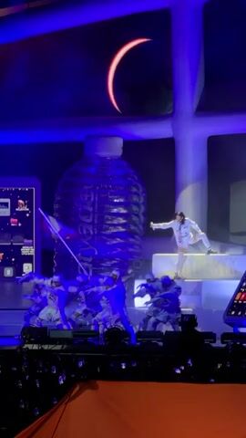 A stage performance is captured from a slightly elevated viewpoint, looking down at the stage. In the background, a large, illuminated screen displays a crescent moon or eclipse. Below the screen, a massive, translucent bottle, appearing to be filled with a blue liquid, dominates the center of the stage. To the left and right of the bottle are large, flat-screen displays showcasing what appear to be app icons.

On the stage itself, a group of performers, dressed in white and blue costumes resembling robotic suits, are positioned in the lower left. They are actively moving, some with arms raised. A Spanish flag is prominently displayed on a pole held by one of these performers. To the right of the group, a female performer, dressed in a white outfit with a pink accent, is seated on a raised platform. She is looking towards the left and appears to be interacting with the scene unfolding below. The stage is lit with vibrant blue and purple lighting, creating a theatrical atmosphere. In the foreground, the heads of an audience are visible, with some holding up phones to record the performance.