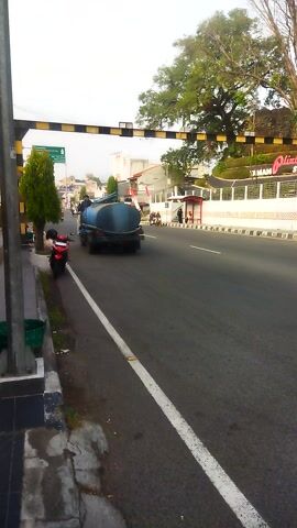 A cement mixer truck is driving on a road next to a sidewalk with a tree and a motorcycle parked on the side of the road. The truck is blue and has a rotating drum. The road is paved and has white lane markings. The sidewalk is made of concrete and has a black and yellow striped pole on the edge. The motorcycle is red and black. The tree is large and has green leaves. The sky is overcast. The scene appears to be in a city or town.