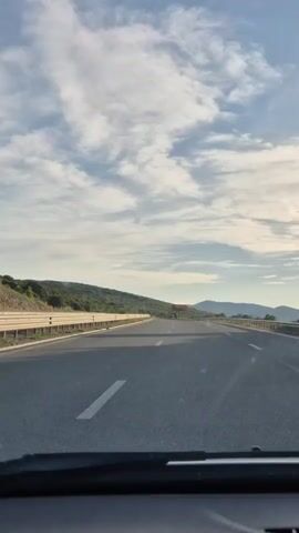 The video shows a view from inside a vehicle moving along a highway. The highway has multiple lanes and is bordered by guardrails. On the left side of the highway, there are hills covered with vegetation. In the distance, there are mountains. The sky is blue with scattered clouds. The sun appears to be setting or rising, casting a warm light on the scene. There is a traffic sign visible in the distance. No people or other vehicles are clearly discernible.