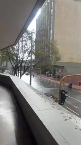 The video shows a view of a city street on a rainy day from a covered walkway. Rain is falling on the street, which appears wet and reflective. Traffic is visible, including cars and a small truck. There are pedestrians on the sidewalk across the street. A tall building is visible in the background. A traffic light is visible on the right side of the frame, showing a green light. The walkway is made of concrete and is partially covered by a white ceiling. A large object is seen being moved across the street.