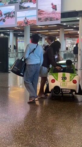 The video shows an indoor shopping mall. In the foreground, a woman wearing a brown shirt and shorts is helping a young boy in a toy car. The toy car is white and green with the number 20 on it. The boy is sitting in the driver's seat, and the woman is reaching into the car, possibly adjusting something or interacting with the boy. Behind them, a woman wearing a light blue sweatshirt and jeans stands facing away from the camera, looking towards the toy car. She is carrying a black bag over her shoulder. Further in the background, other shoppers are visible, and stores are apparent. Large digital screens displaying advertisements are mounted on a wall above the scene. The floor is a speckled terrazzo.
