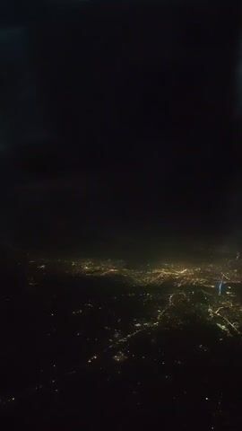 This is a video taken from an airplane at night, showing a city with lights spread out below. A portion of the airplane's wing, which is orange at the tip, is visible in the upper left corner of the frame. The city lights create a textured pattern of yellow and white against the dark landscape.