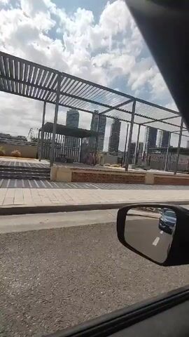 From inside a moving vehicle, the video shows a view of a city street on a partly cloudy day. A covered structure with a metal frame and horizontal slats is visible on the right, positioned over a brick and stone patio. In the background, several tall modern buildings can be seen. The vehicle passes by a pedestrian crossing with a white crosswalk, and a large tree is visible on the right. A person is walking on the sidewalk near a trash can. The side mirror of the vehicle reflects another car behind.