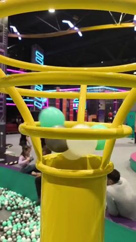 The video is a close-up shot of a yellow tube with several colorful balls inside. The balls are of different colors, including white, green, and gray. The balls are moving inside the tube, and some appear to be suspended in mid-air. In the background, there are blurry figures of people, including a child sitting on a ledge, and a ball pit filled with many small balls. The environment appears to be an indoor recreational facility with bright neon lighting and industrial-style architecture.