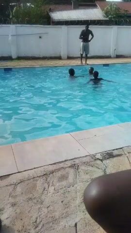 The video depicts an outdoor swimming pool with several people swimming in it. The pool is rectangular and filled with clear blue water. A tiled border surrounds the pool. Some of the swimmers appear to be children. On the pool deck, there is a black shoe, a blue and white striped towel, and various inflatable pool toys. Cars and buildings are visible in the background. The weather appears sunny.