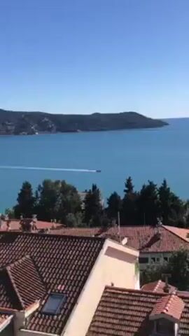 The video offers a scenic panoramic view of a coastal landscape on a clear, sunny day. It begins with a medium shot looking down over the terracotta-tiled roofs of houses. Beyond the houses, a large body of clear blue water stretches out, across which a small white boat with a visible wake is moving from left to right. Dark green trees are scattered among the buildings and along the shoreline.

As the video progresses, the camera slowly zooms out and pans to the right, gradually revealing a broader perspective. The final shot is a wide, sweeping panorama showcasing a large bay or inlet, framed by numerous mountains. Many of the distant mountains have visible snow-capped peaks, indicating either a high altitude or a cooler time of year. The sky is a brilliant, vibrant blue with only a few wispy white clouds scattered near the mountain range. Below the mountains and along the water's edge, more residential areas and what appears to be a small town or village are visible. The hillsides leading down to the water are covered with a mix of lush green and some bare deciduous trees. The overall impression is that of a picturesque and tranquil Mediterranean or Adriatic coastal setting.