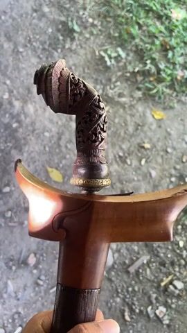 A hand holding a kris. The kris has a wooden handle with intricate carvings and a golden band. The guard of the kris is made of wood and is curved. The background is out of focus and appears to be a dirt or gravel surface with some green foliage. The video is filmed from a high angle, looking down at the kris.