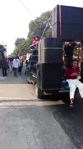 A pickup truck with a flatbed is driving down a paved street. The truck is loaded with large speakers, some of which are stacked high and secured with green straps. Several people are riding in the truck bed, including at least one child wearing a red shirt sitting on top of the speakers and another person standing near the speakers. A woman in a red shirt and white pants is sitting on the edge of the truck bed, looking at her phone. The street is lined with buildings on the left side, appearing to be shops or stalls. People are walking on the sidewalk on the left side of the street, and there is an Indonesian flag attached to one of the buildings. A motorcycle is visible in the street ahead of the truck. The sky is bright and clear, suggesting it is daytime. The overall scene appears to be a procession or a moving event involving music and people.