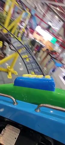 The video shows a person riding an amusement park ride. The camera is mounted low on a blue, green, and yellow car that spins around in a circle as it moves up and down. Other yellow beams fly around the car. The ride is located inside of a building, and parts of the ceiling, lights, and red accents are visible.