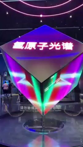 The video shows a rotating exhibit in a museum. The exhibit is a diamond-shaped structure with multiple screens displaying images and text, including the letter H in a circle. The screens are lit with colorful neon lights in shades of green, pink, and white. The structure sits on a mirrored stand and is surrounded by a glass enclosure. There are signs in Chinese and English that read "DO NOT TOUCH THE SCREEN" and "SHOCK HAZARD." In the background, a few people are visible near other exhibits. Overhead, a neon display of concentric circles resembles planetary orbits.