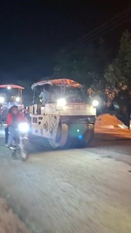 The video shows an asphalt paving operation at night. Several workers are visible, some wearing high-visibility vests. One person is operating a large asphalt roller, which is compacting the freshly laid asphalt. Another worker is using a broom to clear debris from the road surface. In the background, another piece of paving equipment, possibly an asphalt paver, is also active, with bright lights illuminating the work area. Trees and power lines are visible in the background, indicating the work is being done on a street. The overall scene depicts a busy and active construction site under artificial lighting.