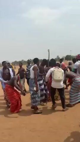 A group of at least 15 men are dancing in a circle on a dusty, brown, sandy field. Some of the men are wearing traditional wraparound skirts in various colors and patterns, some are bare-chested, and others have shirts. One man is carrying a staff. The background features scattered trees and a hazy sky. The video appears to be shot outdoors in daylight.