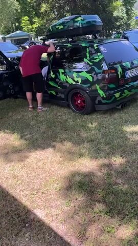 The video shows a person with dark hair wearing a maroon t-shirt and black shorts, standing next to a car with a camouflage wrap and a roof box. The person is leaning into the open driver's side door, appearing to adjust something inside the car. Another person, wearing a white t-shirt, is visible inside the car. The car has red rims and is parked on a grassy area. The background includes trees and other vehicles.