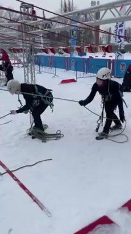 The video shows two people navigating a rope obstacle course in the snow. They are both wearing helmets, harnesses, and dark clothing. They are standing on snowshoes and using ropes to pull themselves along the course. The course appears to be a series of overhead cables or ropes connected by metal supports. There are red and blue banners in the background. There are also other people in the background on the course and observing. The sky is overcast.