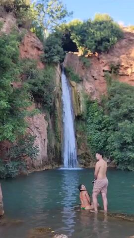 A man and a woman are standing in shallow water in front of a waterfall. The woman is wearing a bikini and is kneeling in the water, facing the waterfall. The man is standing behind her, facing away from the camera, wearing shorts. The waterfall cascades down a rocky cliff face surrounded by green trees. The water in the pool is a clear turquoise color. The sky above is clear and blue.