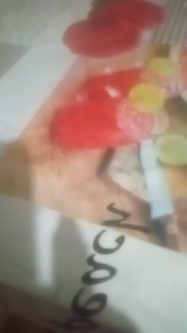 The video shows a close-up, blurry view of a table with a tablecloth that has an image of fruit and drinks. The image features red fruit, possibly strawberries or cherries, and clear glasses filled with red liquid. There are also slices of lime and green herbs. Text that appears to be in black, cursive font is visible on the right side of the tablecloth, reading "beach." The video is shaky and appears to be filmed by someone moving their phone around. There is no discernible action or interaction happening beyond the filming of the table. The lighting is dim.