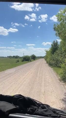 The video shows a point-of-view shot from a vehicle traveling on a gravel road. The road stretches into the distance, bordered by green fields on the left and trees on the right. The sky is blue with scattered white clouds. The front of the vehicle, including what appears to be dark-colored upholstery or fabric, is visible in the foreground. The camera appears to be mounted on the dashboard or a similar interior point of the vehicle. The overall impression is a scenic drive through a rural landscape on a sunny day.