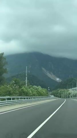 The video shows a road leading through a mountainous landscape under a cloudy sky. The road is asphalt with white lane markings. On the left side of the road, there is a metal guardrail and lush green trees. Further back, a tall electrical pylon stands against the backdrop of a tree-covered mountain. A section of the mountain appears to have a landslide or bare rock face. On the right side of the road, there are streetlights and more trees. The clouds are heavy and grey, suggesting an overcast or possibly rainy day. The perspective is from a vehicle moving forward on the road.