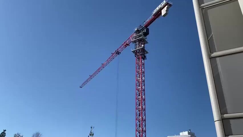 A red tower crane with a long horizontal boom is silhouetted against a clear blue sky. The crane is positioned to the left of the frame, with its tall vertical mast extending upwards. A section of a modern building with a grey, textured facade and white vertical framing is visible on the right side of the frame, partially obscuring the crane. The boom of the crane extends out to the left, creating a strong diagonal line. The scene appears to be an outdoor construction site.