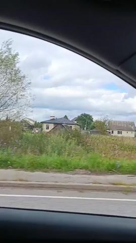 The video shows a view from inside a vehicle, looking out at a rural landscape. The time stamp indicates the footage is from August 2023. The scene features several houses, some appearing older with wooden structures and others more modern. The houses are surrounded by vegetation, including tall grass and trees. A grey fence runs along the foreground, separating the road from the properties. The sky is overcast with clouds. The camera is moving, suggesting the vehicle is in motion. There are no people visible in the frame.